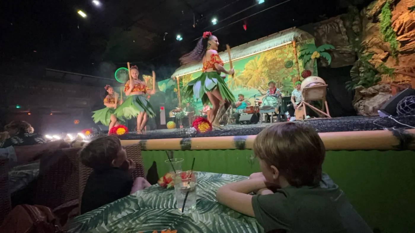 Female hula dancers in green leaf skirts perform with sticks as two young boys watch from their table