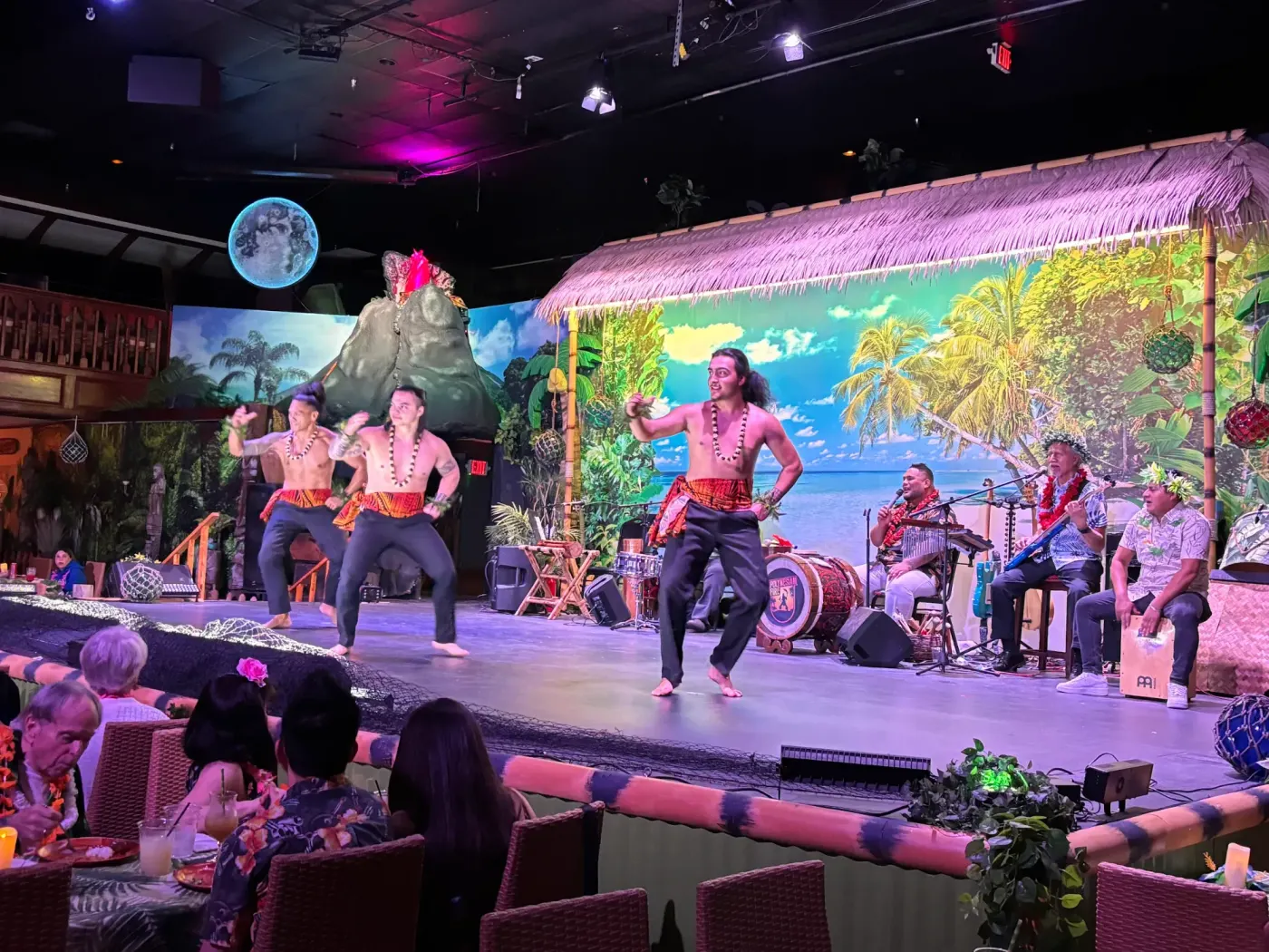 Samoan dancers perform in front of a volcano and beach backdrop with a live band at the Polynesian Fire Luau in Orlando