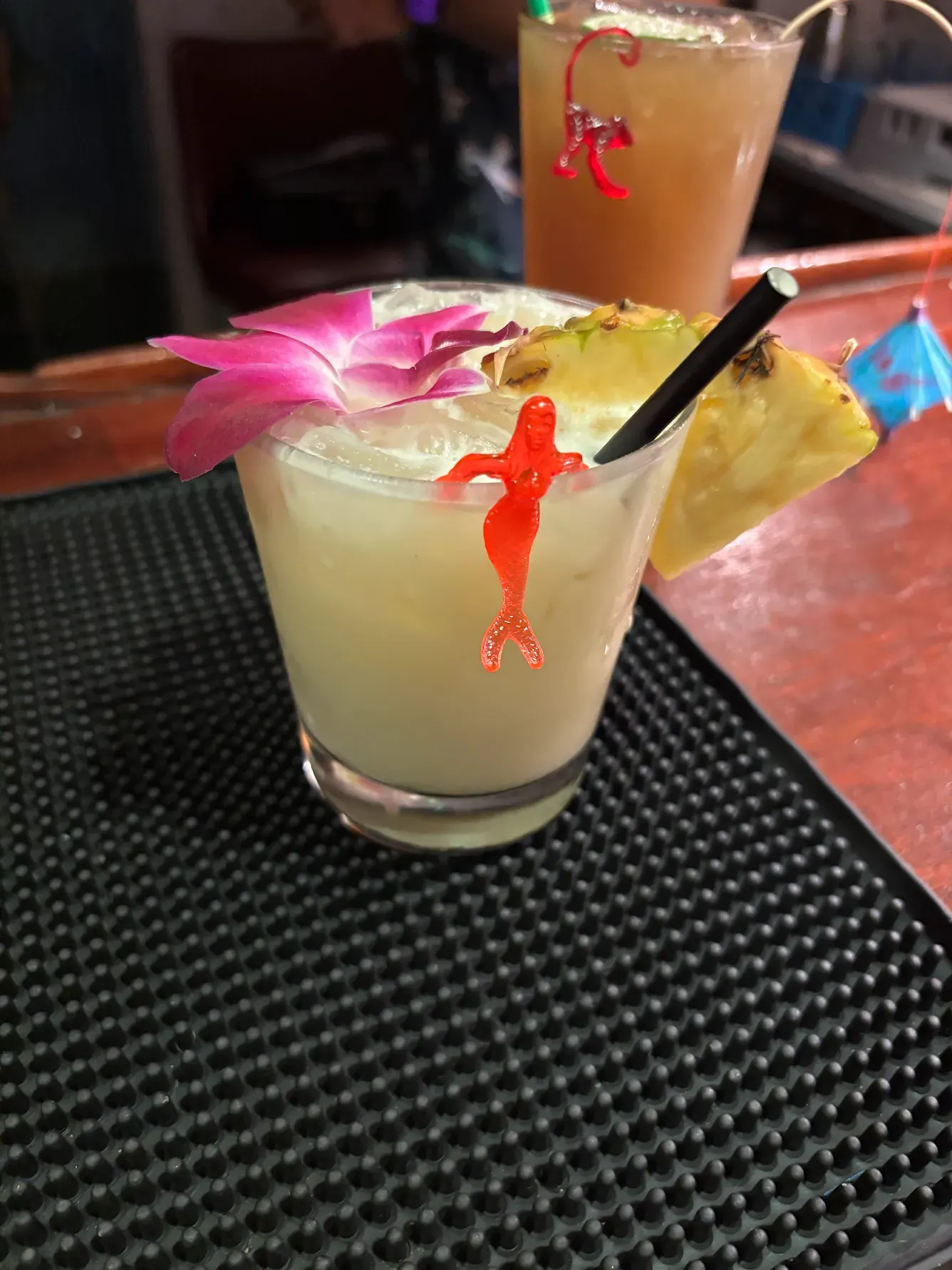 A tropical cocktail with an orchid, pineapple wedge and red mermaid hanging on the glass